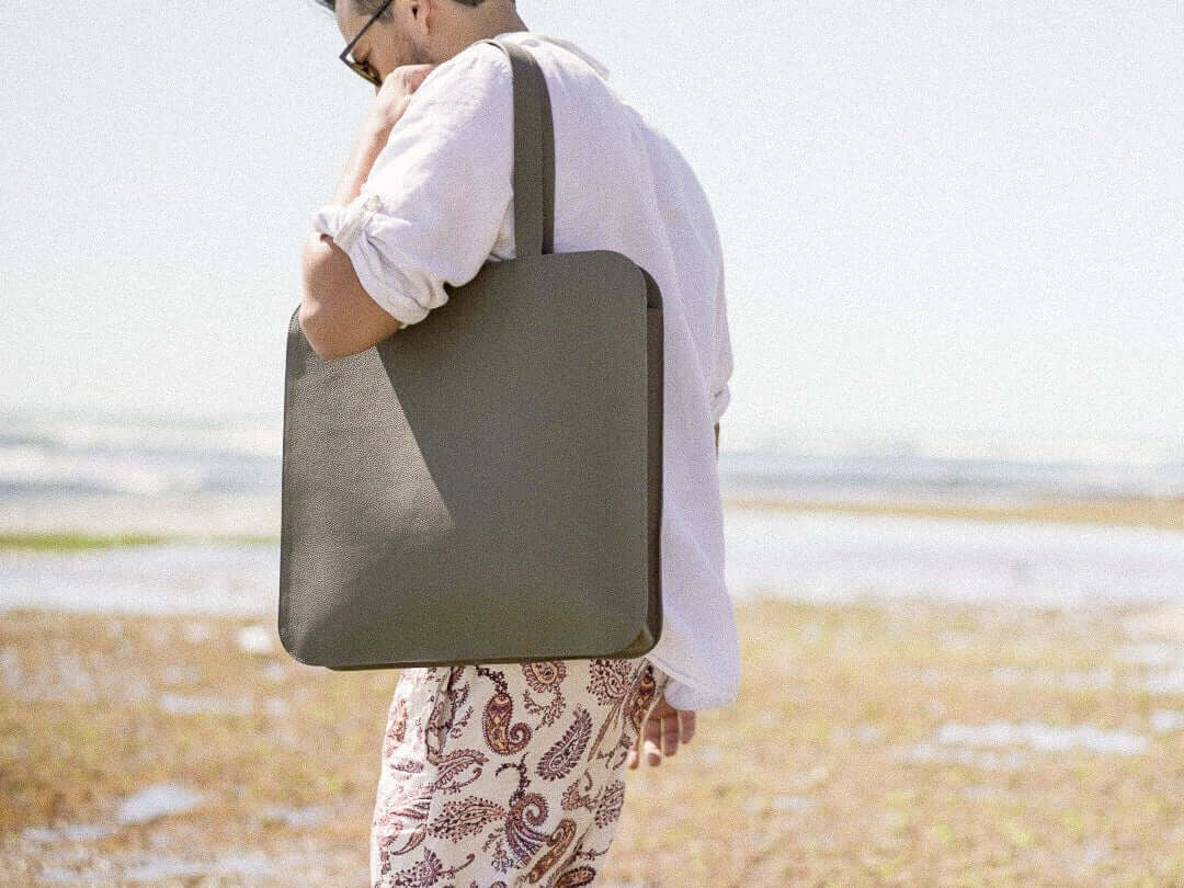 Man carrying handcrafted leather Laptop Tote Bag on a beach, wearing a white shirt and patterned shorts.