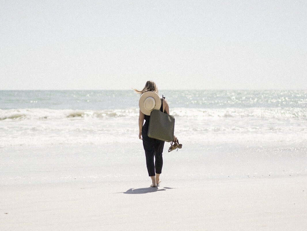 Woman walking on a sandy beach carrying a large tote bag and a sun hat, facing the ocean under a clear sky.